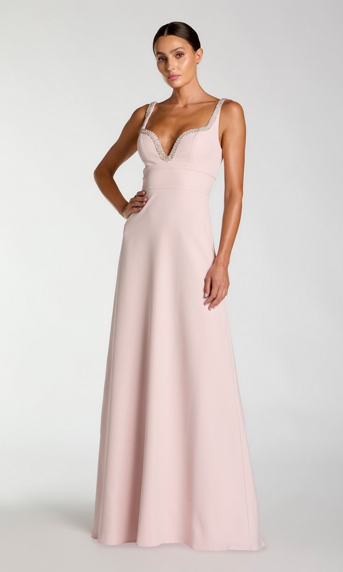 A woman wearing the Mac Duggal 27633 long formal dress in dusty rose, featuring a pale pink crepe fabric with a deep V-neckline and sparkling rhinestone trim, stands against a plain light gray background. This elegant design showcases the signature evening wear style of Mac Duggal.
